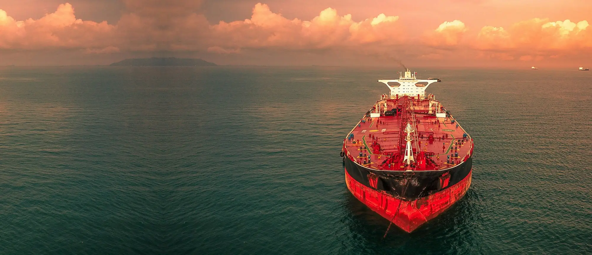 Red and black oil tanker seen head-on on a calm sea under an orange sunset sky